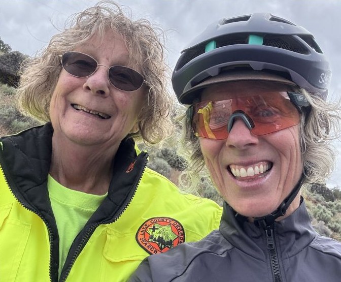 A Lake County Search and Rescue volunteer in a high-visibility jacket takes a selfie with a smiling cyclist in a helmet and sunglasses